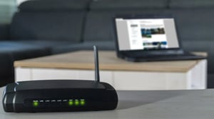 Wi-Fi router with indicator lights on, placed on a table with a laptop in the background.