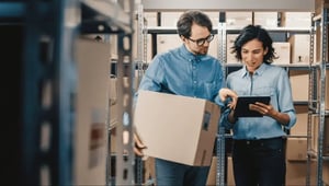 A man holding a box talking to a woman with a tablet, surrounded by shelves with boxes.